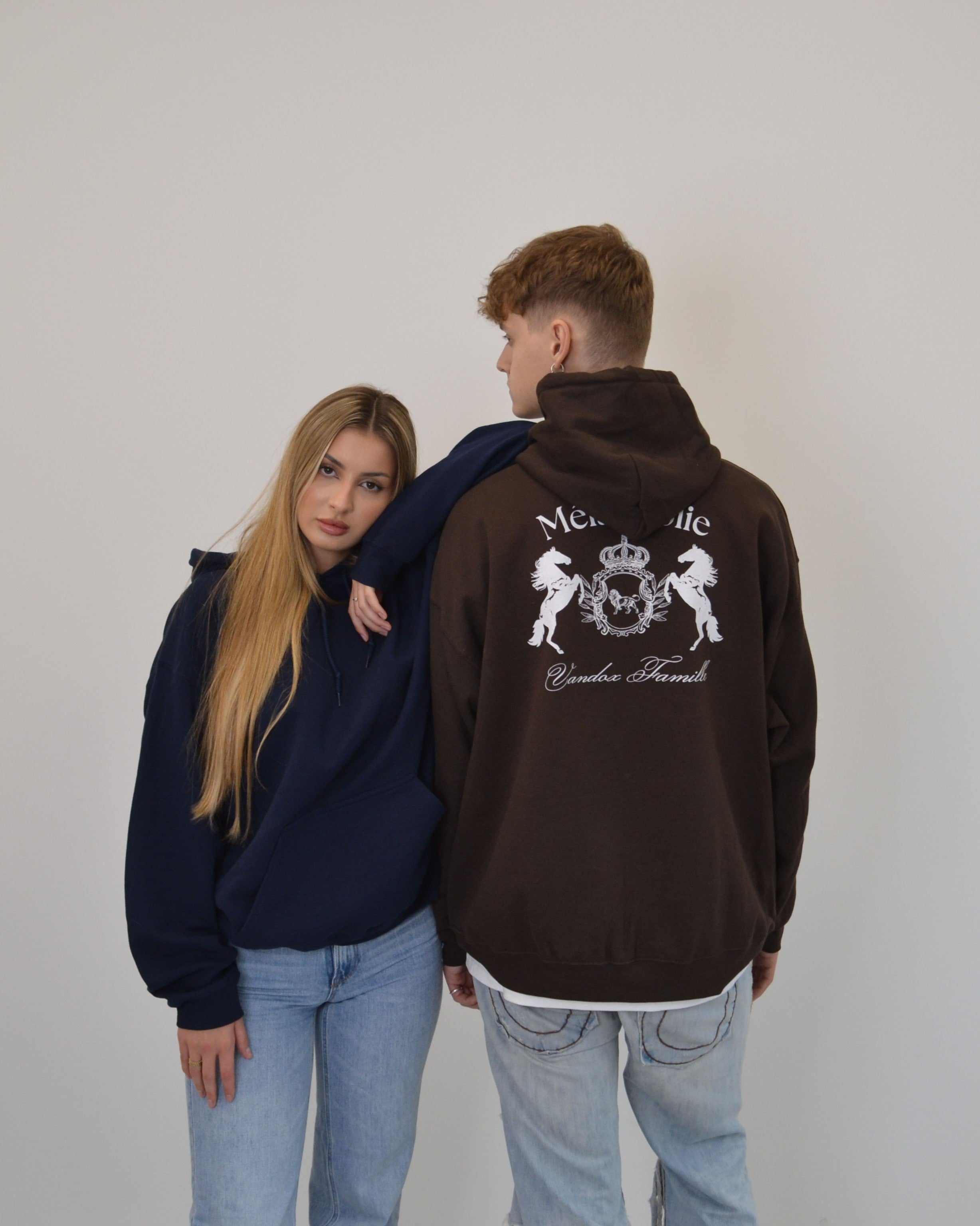 Models wearing Vandox Melancolie Hoodie from the Bestsellers Collection - the woman in navy blue and the man in brown showcasing the detailed back embroidery, standing in a minimal studio setting.