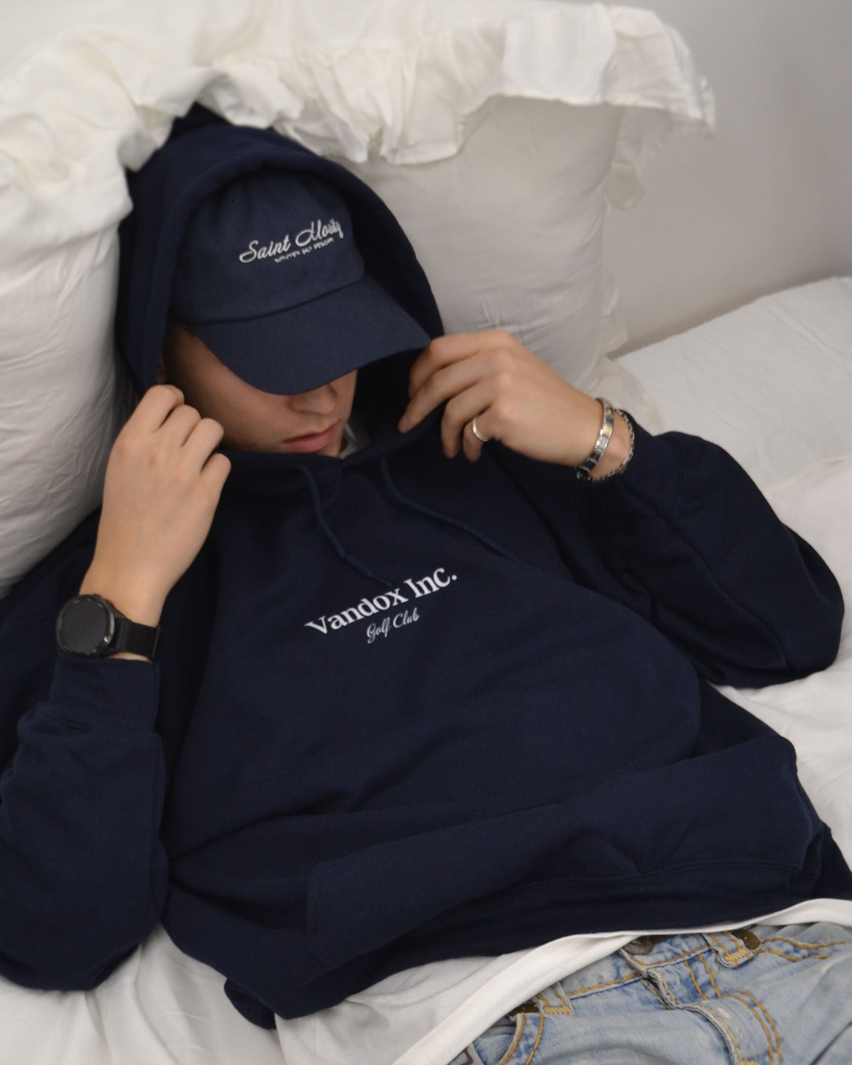 Model wearing the Alpine Escape Cap from the Accessories Collection and Stockholm Hoodie, captured in a relaxed pose on a white bed, showcasing a cozy and refined aesthetic.