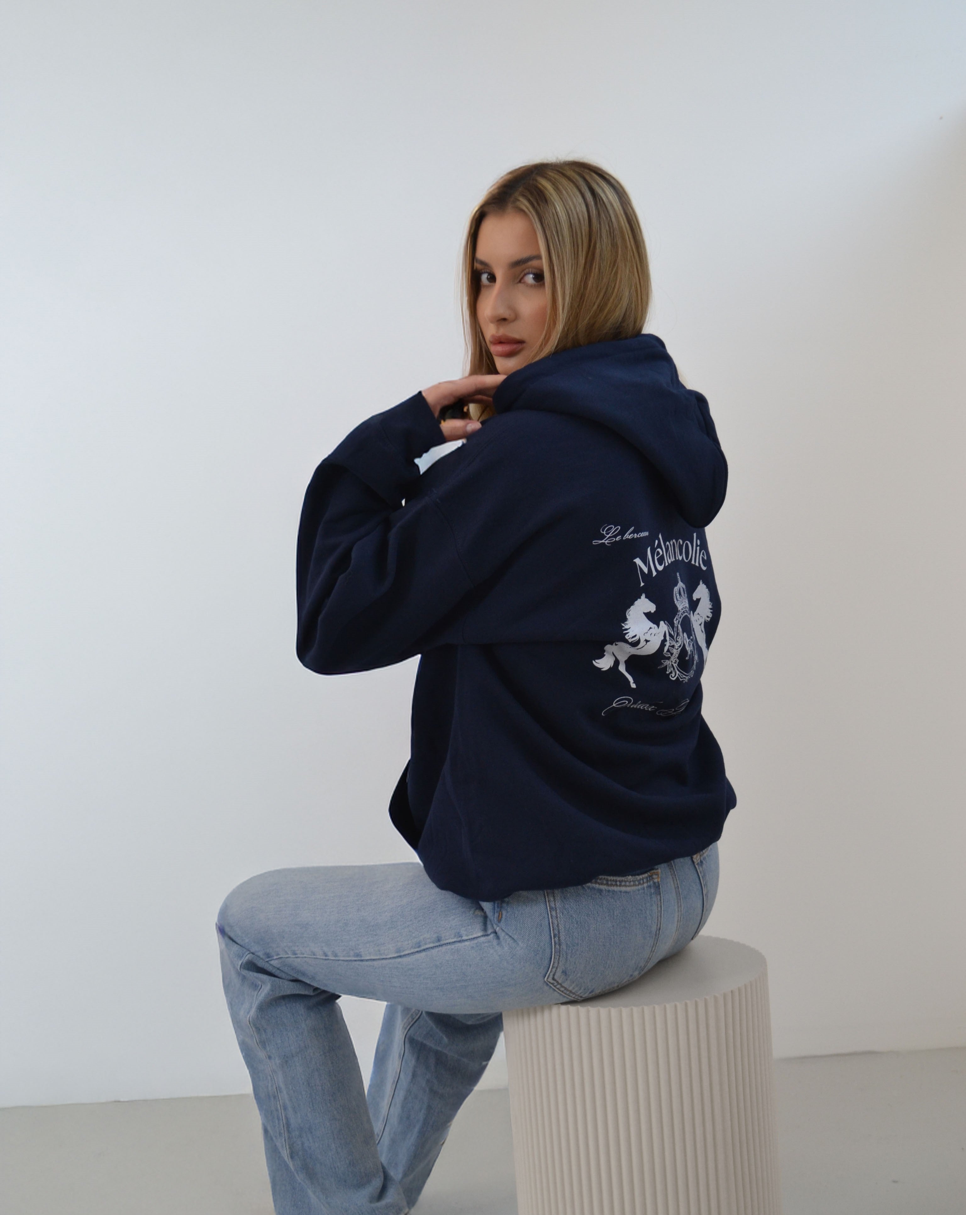 Model wearing the Melancolie Hoodie from the Crewnecks Collection, showcasing the intricate back embroidery while seated in a minimalist studio setting.