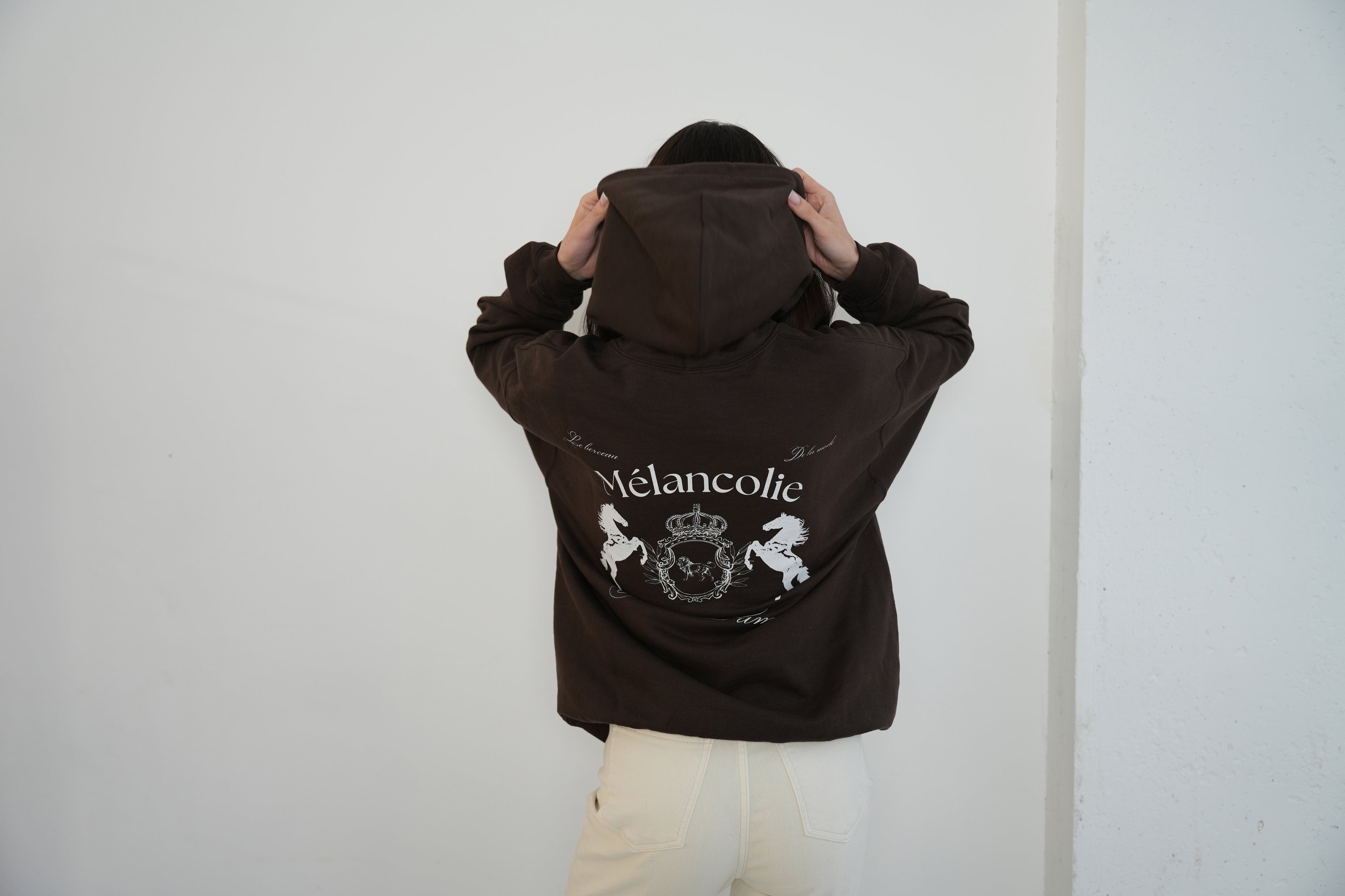 Person wearing a brown hoodie with 'Melancolie' design on the back against a plain background