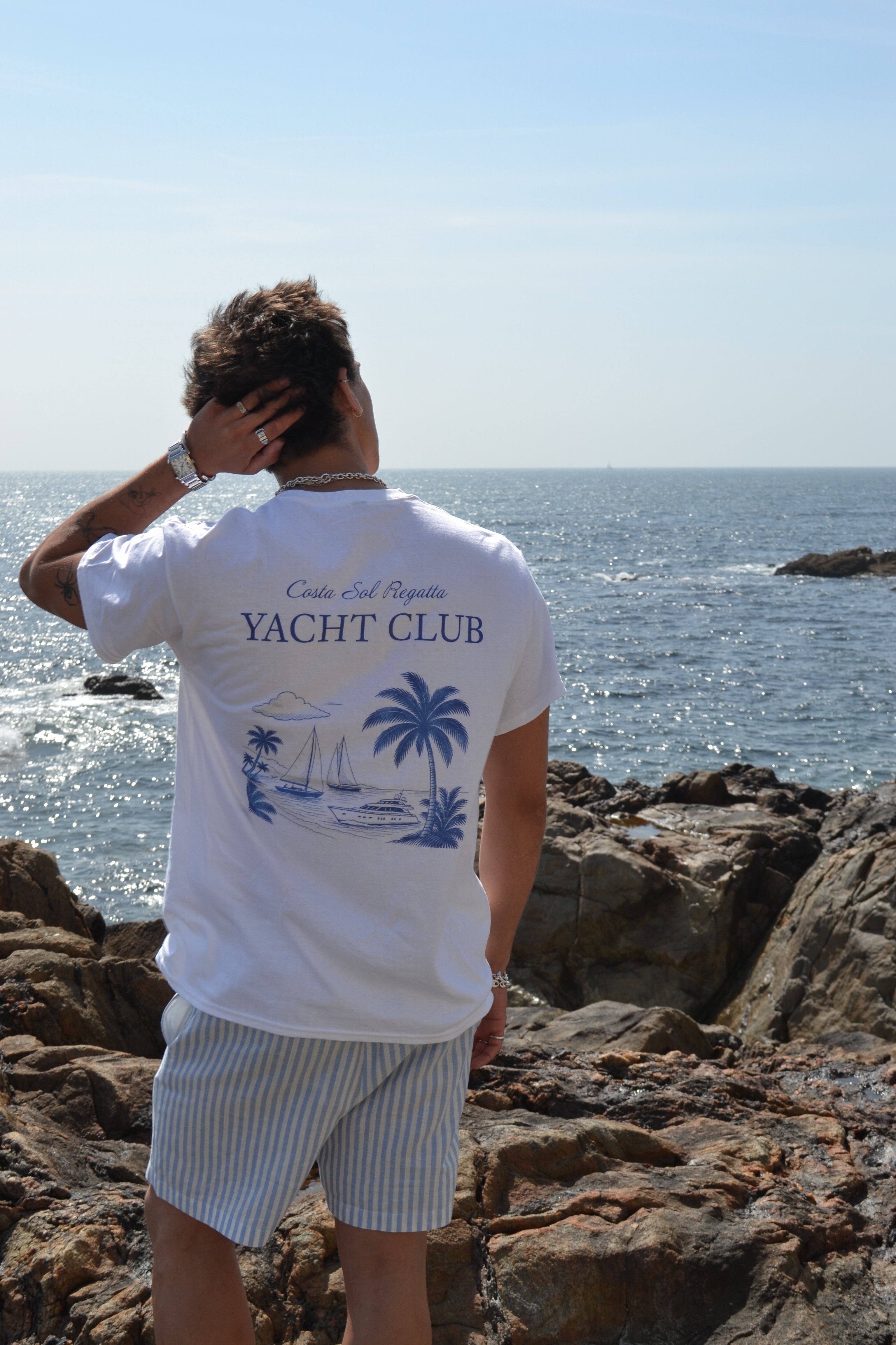 Person wearing a 'Yacht Club' t-shirt with palm tree design by the ocean.
White