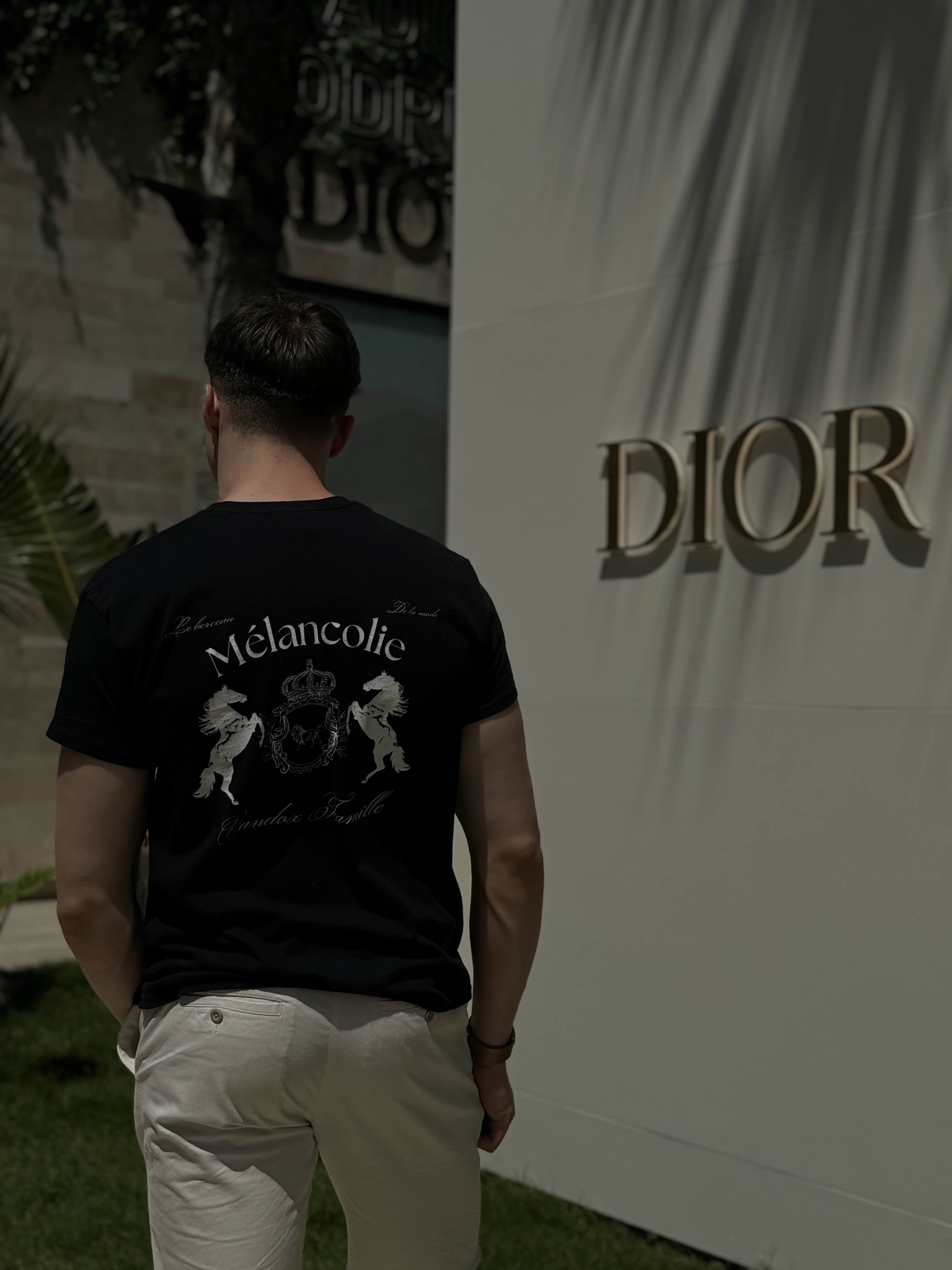 Man wearing the Vandox Mélancolie T-Shirt with crest and horse emblem on the back, standing outside near a Dior store in natural sunlight.