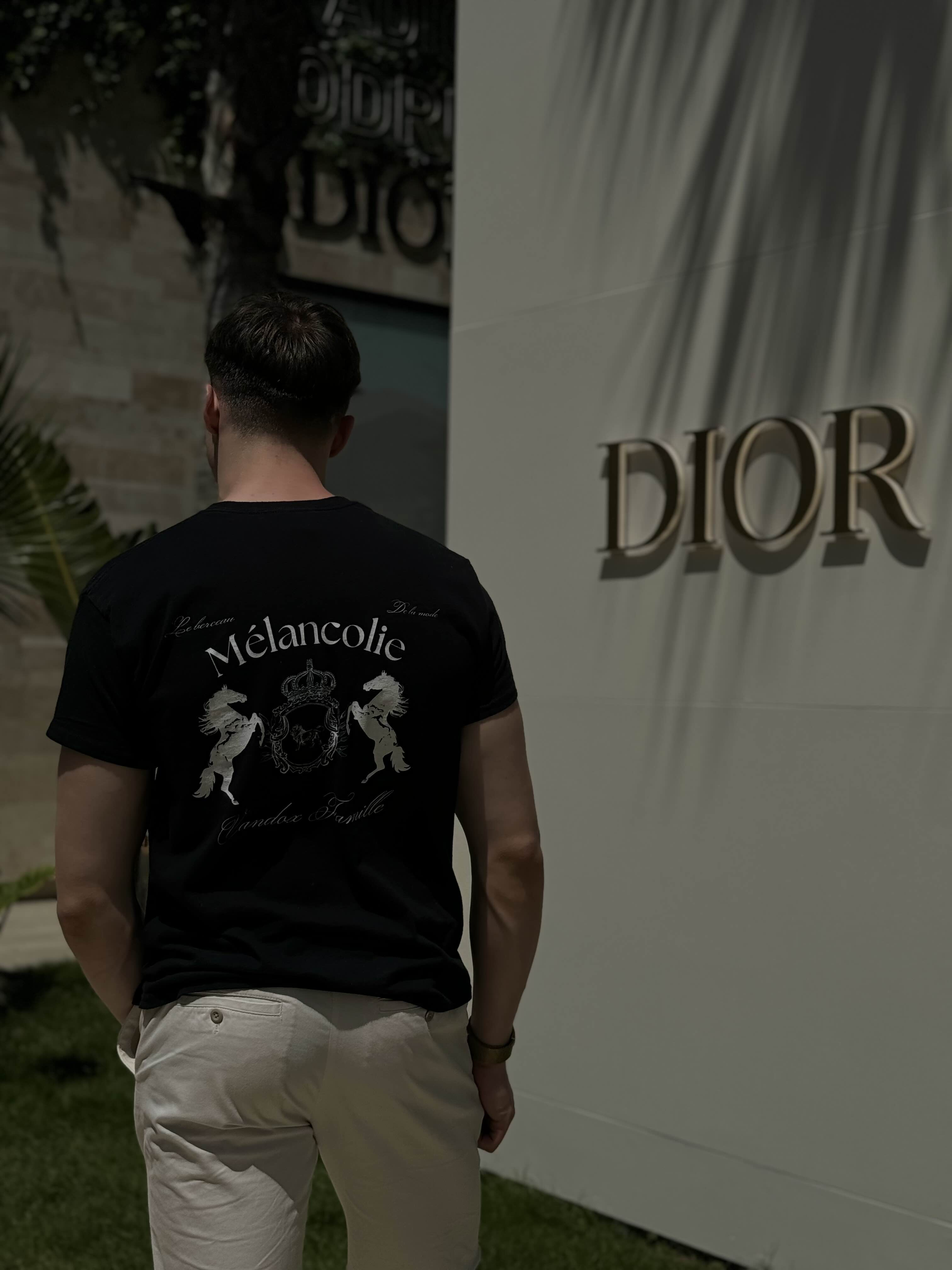 Man wearing the Vandox Mélancolie T-Shirt with crest and horse emblem on the back, standing outside near a Dior store in natural sunlight.