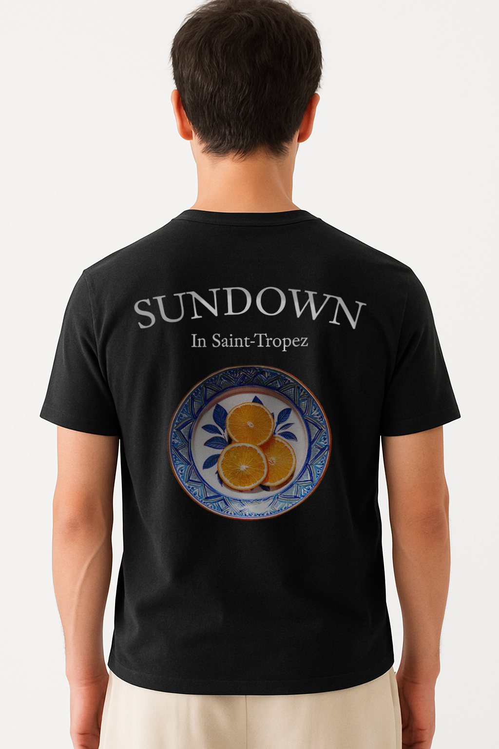 Model wearing the Deep Black Citrus Sunset T-Shirt by Vandox, featuring a hand-painted style plate of oranges with "Sundown in Saint-Tropez" printed above for a bold Riviera-inspired aesthetic.