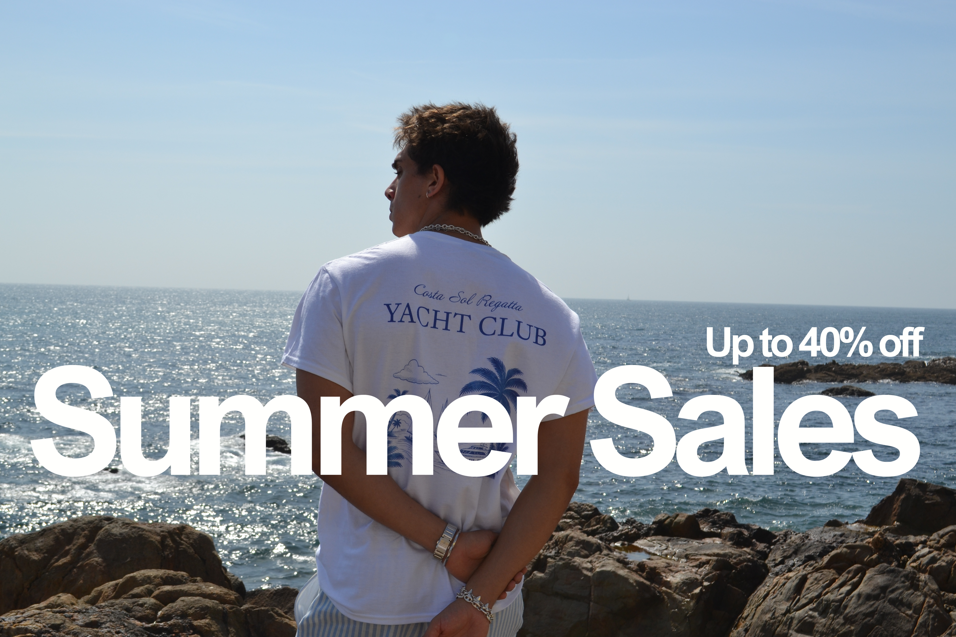 A man stands on rocky shore looking out at the ocean under a clear blue sky, wearing a white vandox riviera regatta T-shirt with 'Yacht Club' printed on the back. Large overlaid text reads 'Summer Sales' and 'Up to 40% off'.
